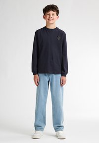 T-shirt a maniche lunghe blu scuro con un piccolo logo bianco, jeans dritti azzurri e sneakers bianche. Design minimalista e vestibilità casual.