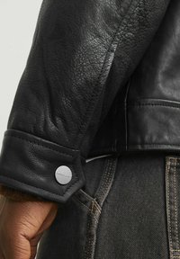 Black leather jacket sleeve with textured finish, featuring a button closure with engraving. Worn over dark denim with visible stitching.