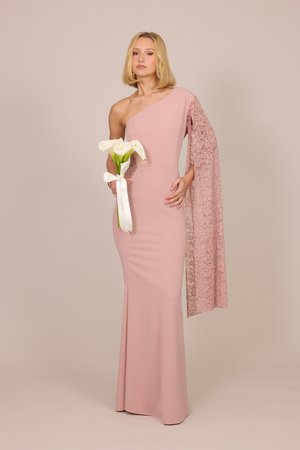 Vrouw in een lange roze jurk met één schouder en kanten mouw die witte callalelies vasthoudt die met een lint zijn gebonden tegen een effen achtergrond.