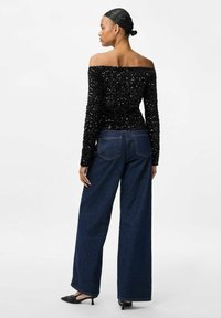 Femme avec les cheveux en chignon portant un haut noir à sequins sans manches, un jean large bleu foncé et des escarpins noirs à bout pointu, se tenant de dos.