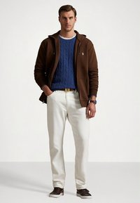 Καφέ φούτερ με φερμουάρ πάνω από μπλε πλεκτό πουλόβερ με ανάγλυφο σχέδιο, λευκό παντελόνι και σκούρα αθλητικά παπούτσια. Φορά ζώνη και ρολόι.