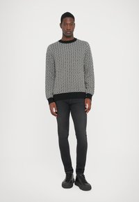 Man standing wearing black and white diamond-patterned sweater, black slim jeans, and black sneakers on white background.