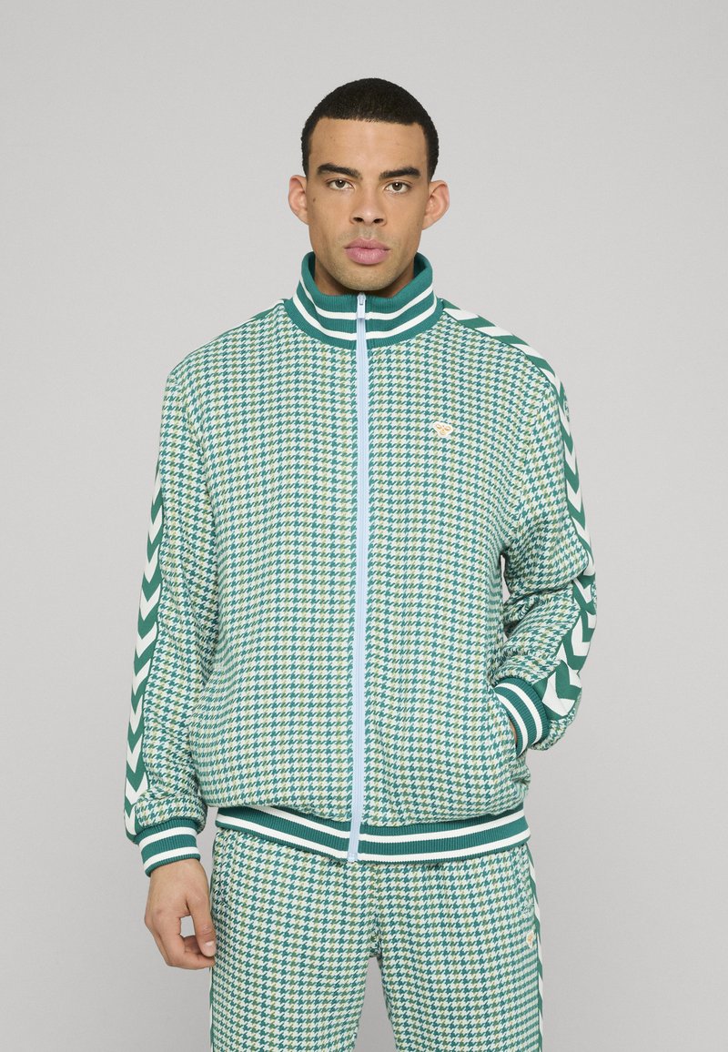 Vihreä-valkoinen houndstooth-kuvioinen vetoketjullinen takki raidallisella kauluksella ja resoreilla, yhdistetty samankaltaisiin housuihin; rinnassa hienovarainen logo.