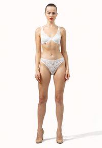 Femme se tenant debout portant un soutien-gorge en dentelle blanche et des culottes taille haute assorties avec des talons nus, sur un fond uni.