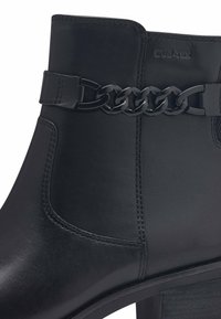 Bottines noires en cuir lisse, avec un détail en chaîne et un talon épais. La chaussure présente une silhouette élégante et des coutures discrètes.