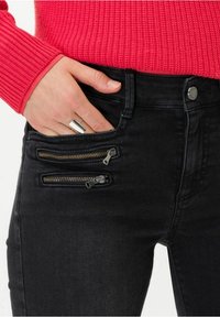 Jeans negros de tiro alto con tres bolsillos con cremallera, hechos de tela de mezclilla. El herraje metálico visible incluye cremalleras doradas y plateadas.