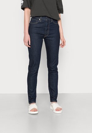 Person wearing dark blue slim-fit jeans, black oversized t-shirt, and white slide sandals standing against a light background.