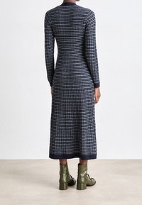 Pletené šaty s dlouhými rukávy, námořnická modř jako základ, bílé horizontální pruhy. Mají přiléhavý střih, dlouhé po kotníky, doplněné zelenými kotníkovými botami.