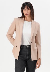 Béžový blazer se střihaným střihem, na jediné knoflíkové zapínání, s klopami a dvěma předními kapsami, přes bílé tričko a tmavé džíny.