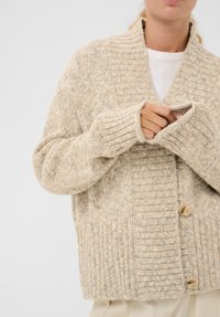 Person, der eine beige Strickjacke mit großen Knöpfen trägt, passt gerade den Ärmel an und kombiniert dies mit einem weißen Oberteil und hellen Hosen.