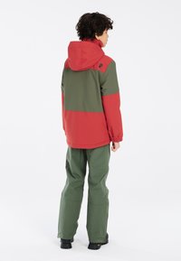 Rote und grüne Jacke mit Kapuze, die ein Farbstreifen-Design aufweist. Kombiniert mit weiten grünen Hosen und schwarzen Stiefeln. Die Textur wirkt glatt.