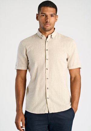 Homme portant une chemise beige à manches courtes et un pantalon foncé, se tenant avec une main dans la poche devant un fond uni.