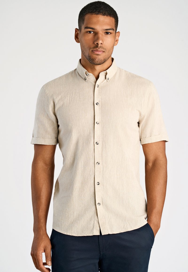 Man in een lichtbeige overhemd met korte mouwen en donkere broek, staand met één hand in zijn zak tegen een eenvoudige achtergrond.