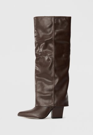 Brown leather knee-high boot with a pointed toe, slouchy design, and chunky square heel. Smooth texture with subtle creasing details.