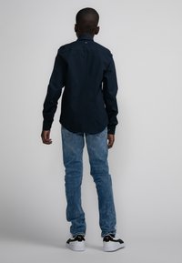 Marineblauwe overhemd met lange mouwen, casual pasvorm, met knoopsluiting; gecombineerd met lichtblauwe jeans en zwart-witte sneakers, gezien vanuit de achterkant.