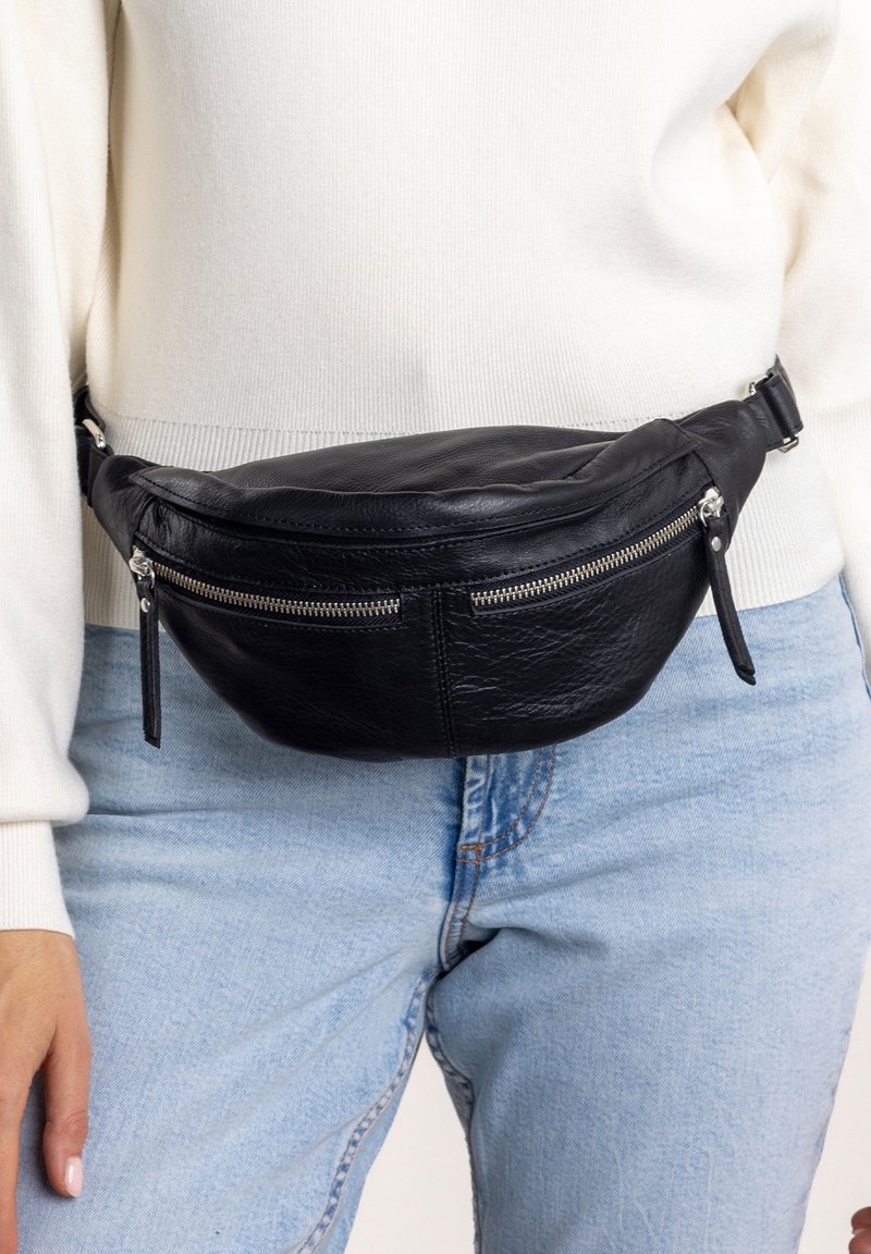 Banane en cuir noir avec deux poches zippées, forme incurvée et finition texturée, portée sur un jean en denim clair.