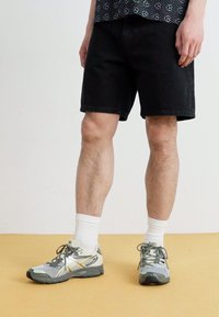 Sorte denim shorts med lige snit, parret med grå og hvide sportssko og hvide ankelstrømper, på et gult gulv.