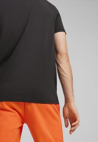 Chemise noire à manches courtes avec un col rond, associée à un pantalon orange vif ; une texture lisse et des coutures nettes sont visibles.