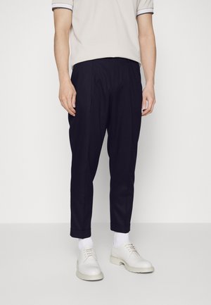 Homme portant un pantalon bleu marine fuselé, des chaussettes blanches, des chaussures blanches à lacets et une chemise blanche à manches courtes avec un liseré noir.