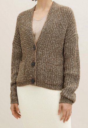 Frau trägt eine braune, grob gestrickte Strickjacke mit Taschen und schwarzen Knöpfen über einem beigen Oberteil und einem weißen, gerippten Rock.