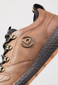 Chaussure en cuir marron avec des œillets métalliques, des lacets noirs, un tissu texturé et une semelle en caoutchouc ornée d'un emblème de marque notable.