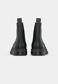 Melnas ādas potīšu zābaki ar gludu virsmu, elastīgām sānu daļām un masīvu gumijas zoli. Aizmugures skats ar minimālistisku dizainu.