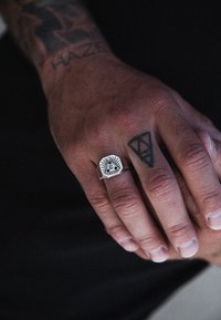 Anello signet argento con design di un occhio circondato da raggi, incastonato su una superficie piatta e texturizzata. Mano con tatuaggi visibili, tonalità della pelle sembra abbronzata.