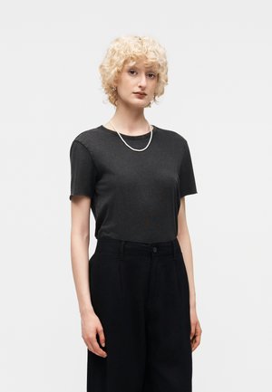 Ξανθό άτομο με κατσαρά μαλλιά, που φοράει απλό μαύρο κοντομάνικο, μαύρο παντελόνι με ψηλή μέση και ένα απλό λευκό κολιέ, στέκεται μπροστά σε λευκό φόντο.