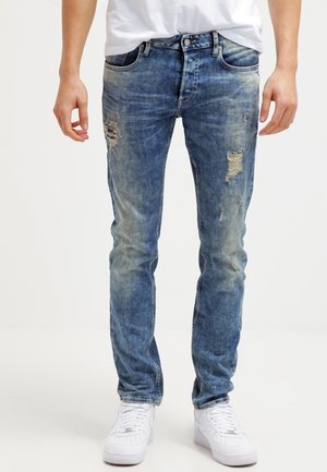 Henkilö, joka pukeutuu slim fit -mallisiin, haalistuneen sinisiin kulutettuuihin farkkuihin, joissa on repeämiä reisissä, yhdistettynä valkoisiin lenkkareihin ja valkoiseen paitaan.