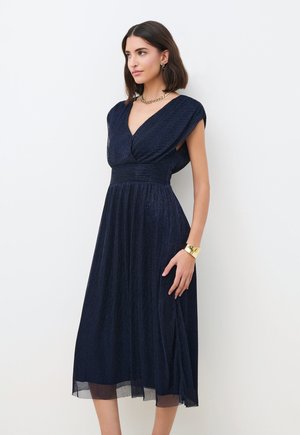 Kobieta ubrana w granatową, bezrękawnikową sukienkę midi z dekoltem w kształcie litery V i plisowaną spódnicą, złoty naszyjnik z łańcuszkiem, kolczyki i złota bransoletka mankietowa.
