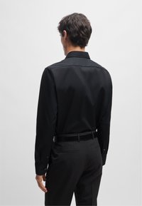 Camicia nera a maniche lunghe in tessuto morbido, caratterizzata da un colletto classico e dettagli con bottoni, indossata con pantaloni scuri e una cintura nera.