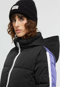 Schwarze Pufferjacke mit lila und weißen seitlichen Akzenten. Hoher Kragen, Reißverschluss und enge Passform. Getragen mit einer schwarzen, gestrickten Mütze.