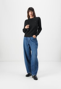 Pull en maille texturée noir avec col rond, porté avec un jean en denim bleu ample. Le modèle tient un sac noir, debout sur un fond blanc.