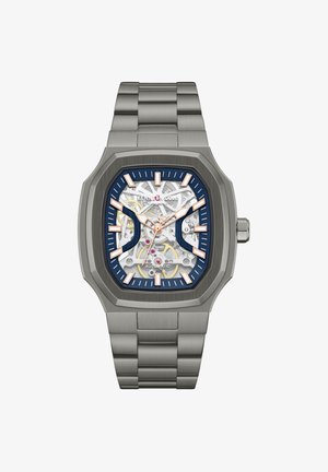 Kvadratna avtomatska zapestna ura iz nerjavečega jekla z vidnimi zobniki, markerji v rožnato zlati barvi in modrimi poudarki na številčnici.