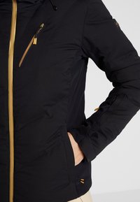 Svart isolerad jacka med guldzipper, sidofickor och en extra ärmficka. Slät textur med kontrasterande detaljer i metall.