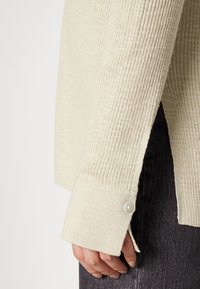 Ljus beige stickad tröja med ribbat mönster, försedd med en knappslutning vid ärmen och en sidslits, ihop med mörka byxor.