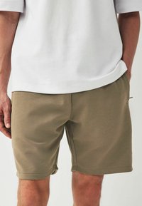 Shorts kaki avec une texture lisse, des poches latérales et des ourlets retroussés, associés à une chemise blanche à coupe ample.