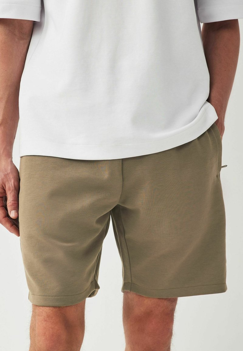 Shorts kaki avec une texture lisse, des poches latérales et des ourlets retroussés, associés à une chemise blanche à coupe ample.