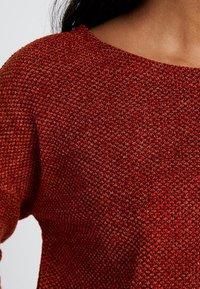 Pull rouge en maille avec un motif texturé, doté d'un large décolleté et d'un matériau doux et flexible. Vue en gros plan du détail du tissu.