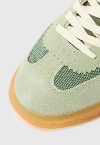 Grønn semsket sneakers med nettingdetaljer, rund tå, kremfargede snørebånd og en gjennomsiktig gummisåle. Teksturert overdel med bølget detaljer.
