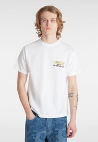 Bílý bavlněný tričko s potiskem žlutého a černého loga "VANS" na levém prsu, volný střih, krátké rukávy. V kombinaci s modrými maskáčovými džíny.