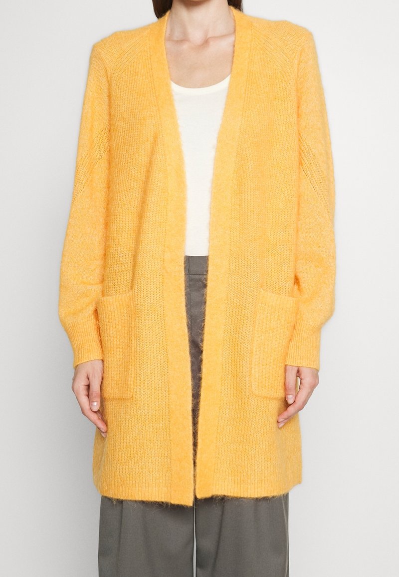 Femme portant un long cardigan tricoté jaune pâle, ouvert sur le devant avec des poches avant, associé à un haut blanc et un pantalon gris.