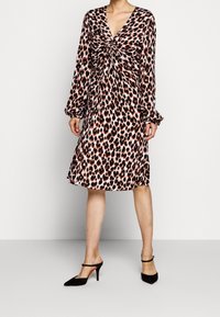 Kobieta w sukience w efekcie panterki o długości do kolan, z długimi bufkami na rękawach oraz czarnych szpilkach na odkrytej pięcie.