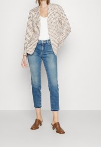 Blazer con estampado en beige claro con diseños geométricos, combinado con jeans azules ajustados y zuecos marrones con tacón redondeado.
