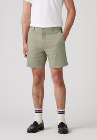 Pantaloni scurți verzi până la genunchi, confec�ționați dintr-un material ușor, cu un front plat și buzunare laterale, asociați cu șosete albe și mocasini negri.