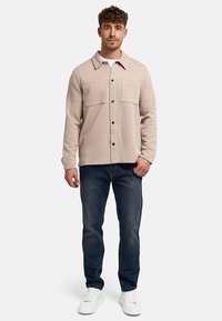 Hombre con camisa beige acolchada abotonada, jeans azules y zapatillas blancas, de pie frente a un fondo liso.