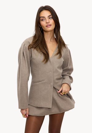 Vrouw met lang bruin haar, gekleed in een taupe blazer met pofmouwen en een bijpassende rok, geaccessoriseerd met gouden ringen en oorbellen in de vorm van een ring.