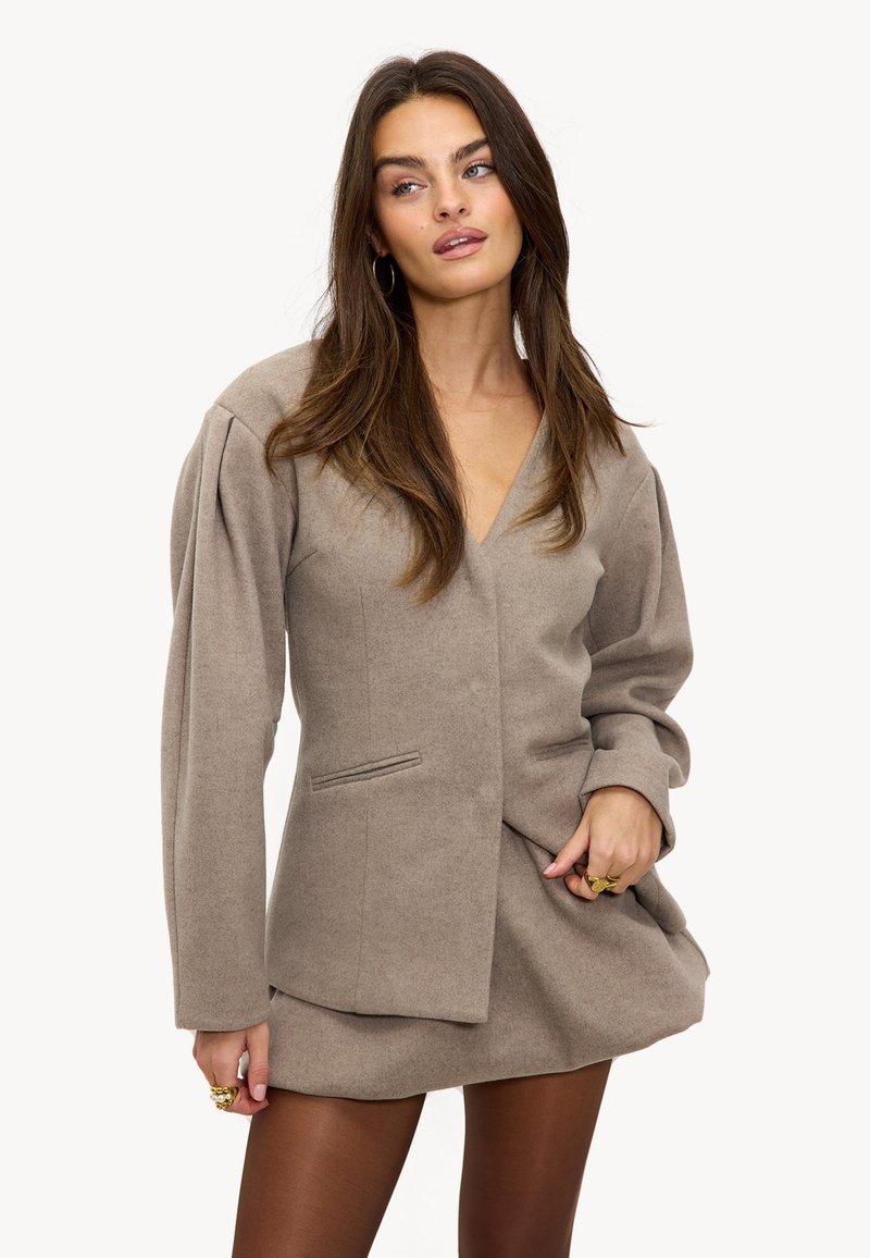 Femme aux longs cheveux bruns portant un blazer taupe à manches bouffantes et une jupe assortie, accessoirisée de bagues en or et de boucles d'oreilles créoles.