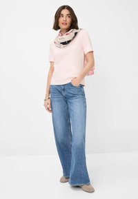 Top rosa chiaro a maniche corte abbinato a jeans a gamba larga blu. Una sciarpa a motivi crema e rosa si drappeggia attorno al collo. Scarpe grigie completano il look.