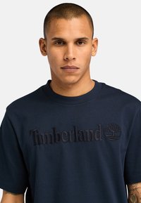 Tyrkysové bavlněné tričko s kulatým výstřihem, na přední straně s vyraženým logem "Timberland" a symbolem stromu. Krátké rukávy.